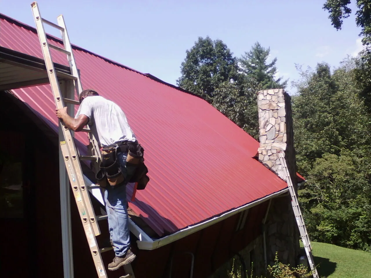Roofer on a ladder beside a red metal roof and stone chimney — Soffit & Fascia Repair by Roofing Brothers in Langley